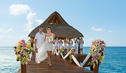 A newly married couple holding hands exiting their destination wedding ceremony held on a tropical hut over bright blue ocean water