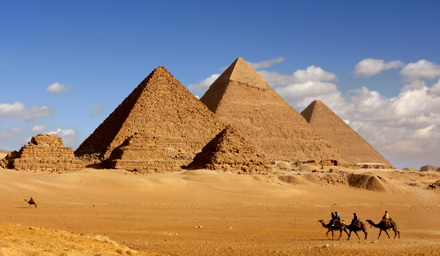 The ancient pyramids set against a blue sky with fluffy clouds and people riding camels in the foreground