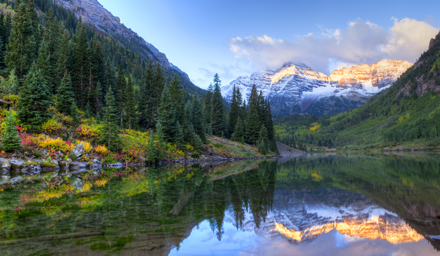 Large snow capped mountain in the background with lush evergreen trees and still water in the foreground